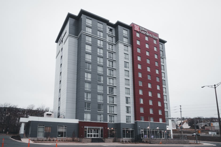 A modern high-rise building with a predominantly gray facade, accented by a vertical red section displaying the Hilton Garden Inn logo near the top. The lower part of the building features large glass